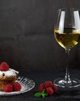 A stemmed wine glass filled with white wine stands on a dark surface beside a small dessert cupcake on a clear plate, with raspberries and mint leaves scattered nearby against a dark textured background.