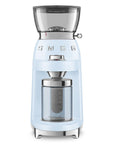 A light blue SMEG coffee grinder with a clear bean hopper, silver metal components, and a clear collection container, shown on a solid white background.