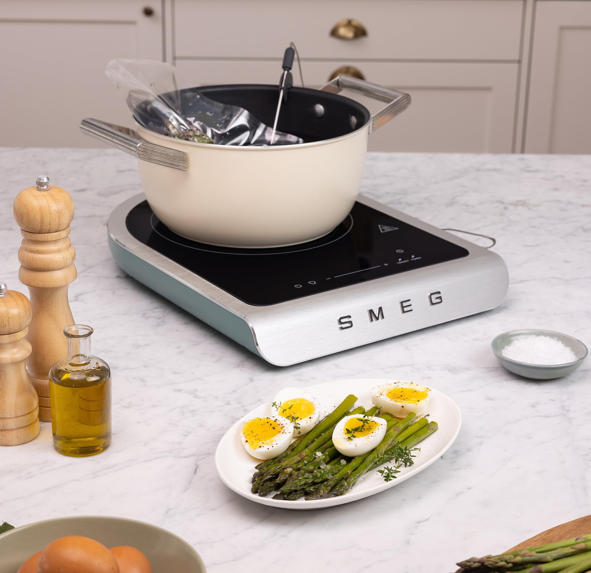 Induction cooktop with a pot on a kitchen counter, featuring a plate of eggs and asparagus.