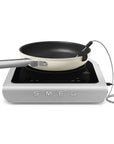 Portable induction cooktop with a frying pan on a white background, featuring the Smeg brand.