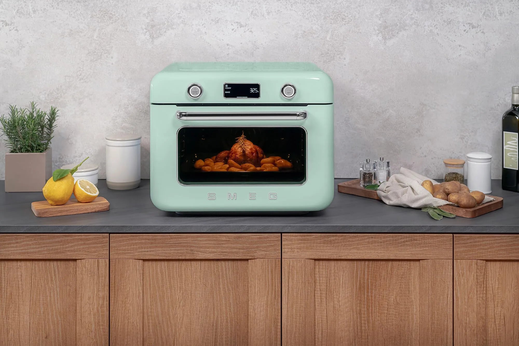 Pastel green Smeg retro line countertop air fry oven on a kitchen counter with food inside, surrounded by kitchen items.