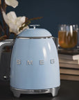Light blue Smeg kettle on a dark surface with books and a glass of tea in the background.