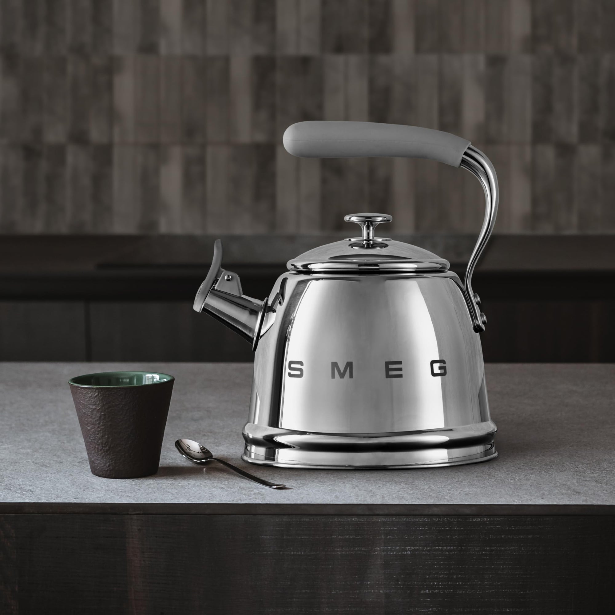 Stainless steel kettle with gray handle on a kitchen counter, featuring the Smeg brand, next to a mug and spoon.