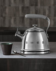 Stainless steel kettle with gray handle on a kitchen counter, featuring the Smeg brand, next to a mug and spoon.