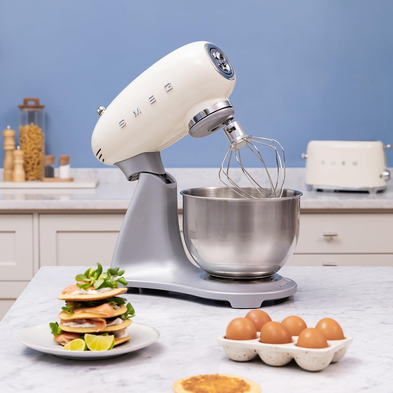 Smeg stand mixer with whisk attachment on a kitchen counter, surrounded by eggs and a stack of pancakes.