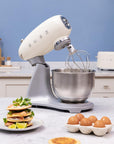 Smeg stand mixer with whisk attachment on a kitchen counter, surrounded by eggs and a stack of pancakes.