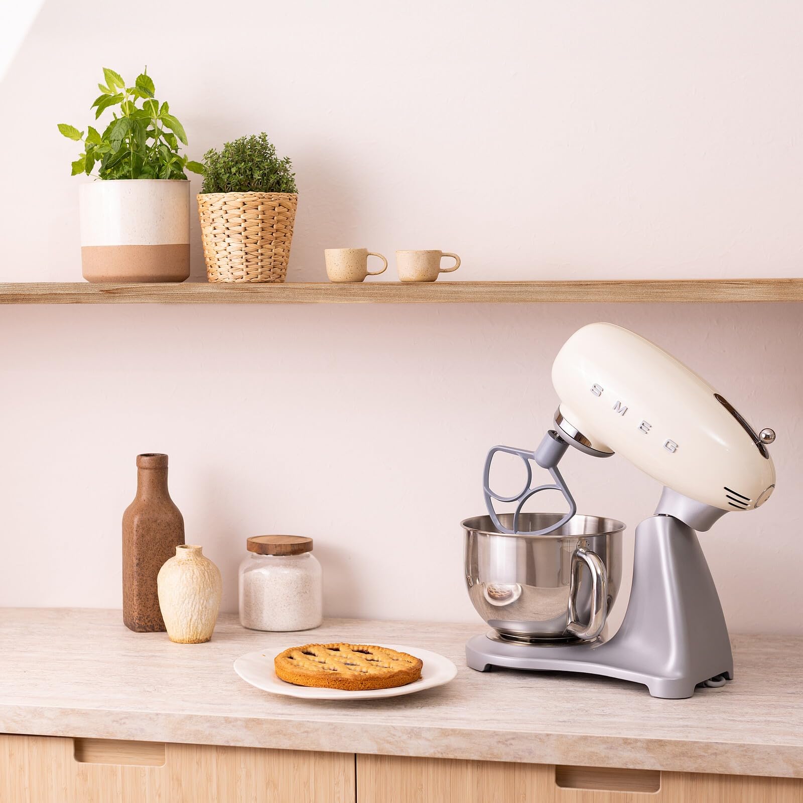 Kitchen scene with a cream Smeg stand mixer on a countertop, pie on a plate, and plants on a shelf.
