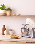 Kitchen scene with a cream Smeg stand mixer on a countertop, pie on a plate, and plants on a shelf.
