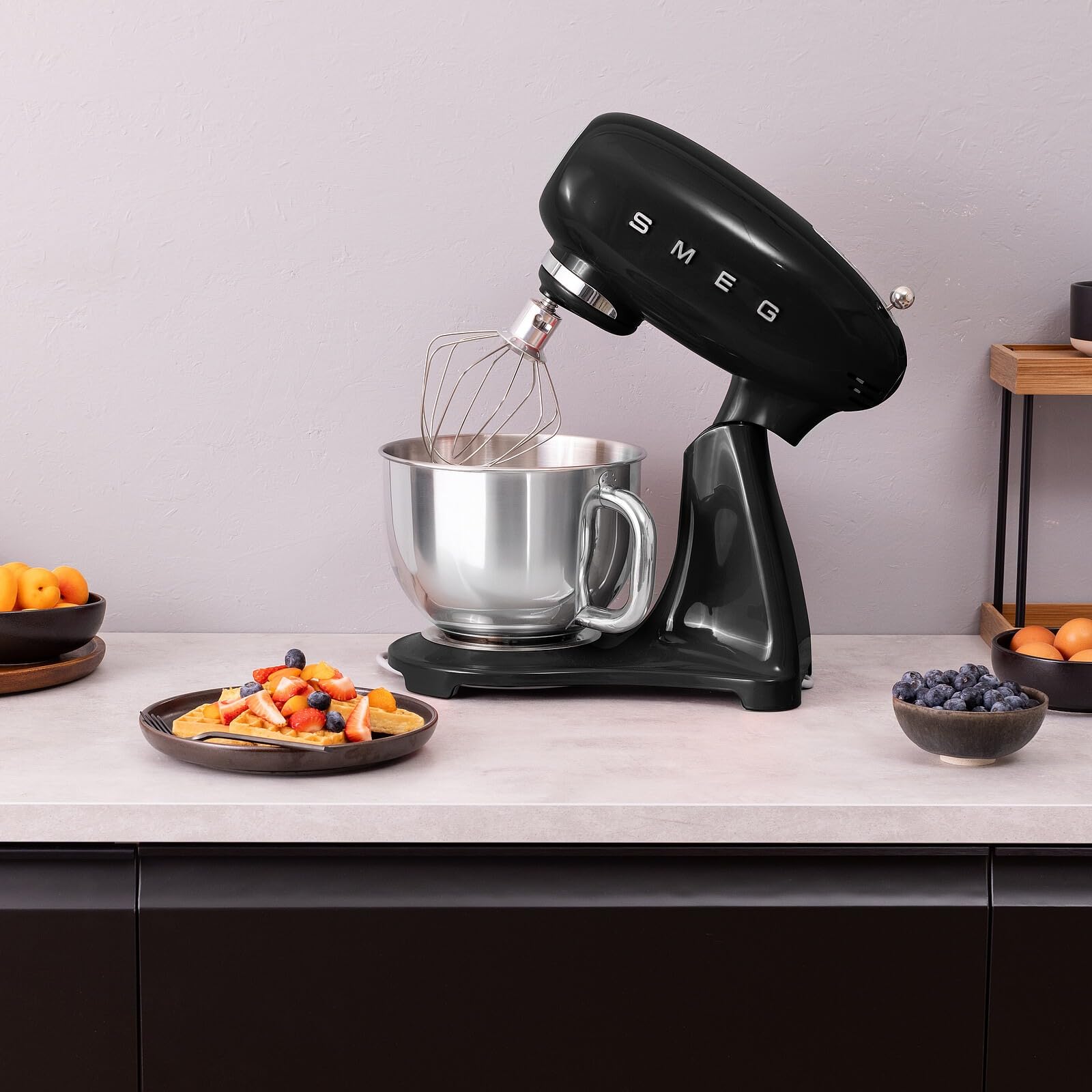 Black Smeg stand mixer on a kitchen counter with fruit bowls in the background