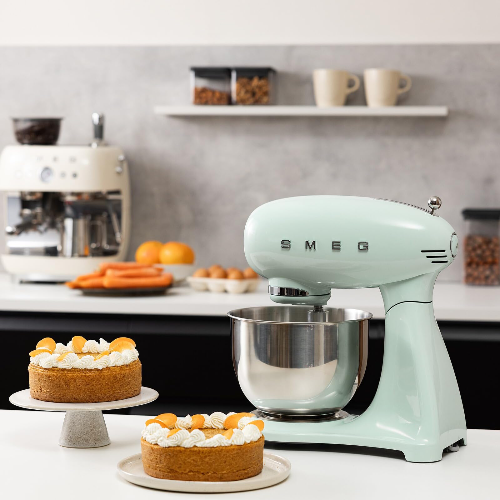 Light green Smeg stand mixer in a kitchen with cakes and fruits on a counter.