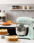 Light green Smeg stand mixer in a kitchen with cakes and fruits on a counter.