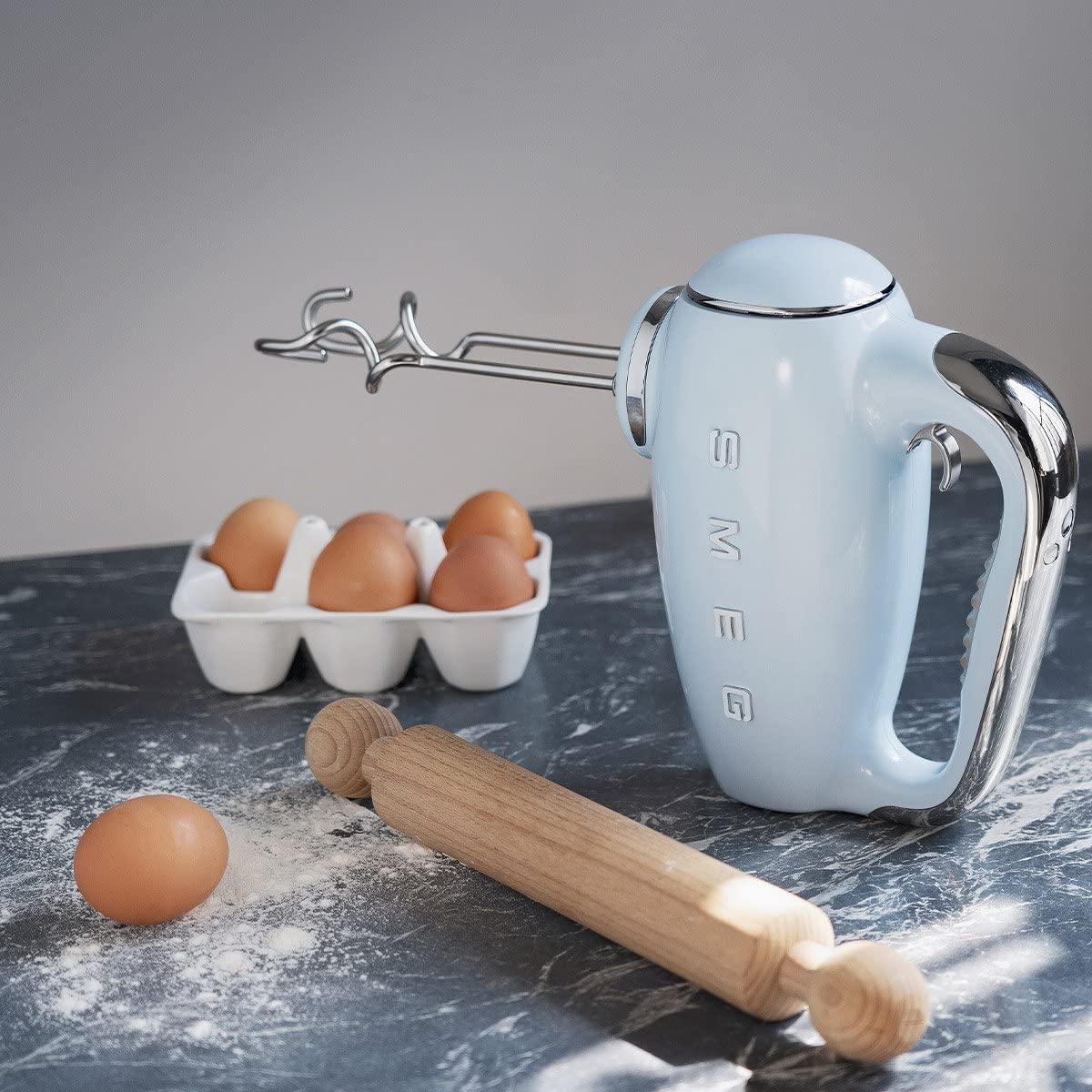 Pastel blue Smeg hand mixer next to baking materials on a black marble surface