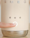 Close-up of a cream milk frother with 'SMEG' branding and control buttons with someone's finger pressing a button on it