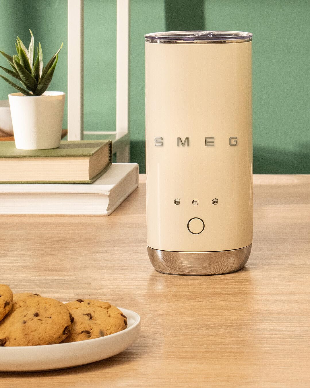 Cream-colored Smeg milk frother on a wooden table with cookies and a plant in the background