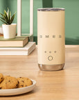 Cream-colored Smeg milk frother on a wooden table with cookies and a plant in the background
