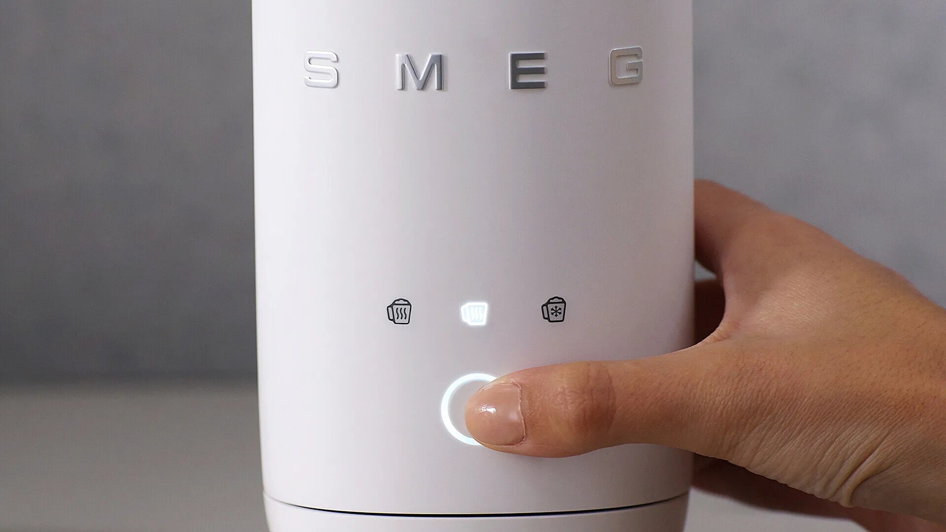 A close-up of a hand pressing the button on a white SMEG milk frother, with the brand name 'SMEG' visible on the front.