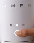A close-up of a hand pressing the button on a white SMEG milk frother, with the brand name 'SMEG' visible on the front.