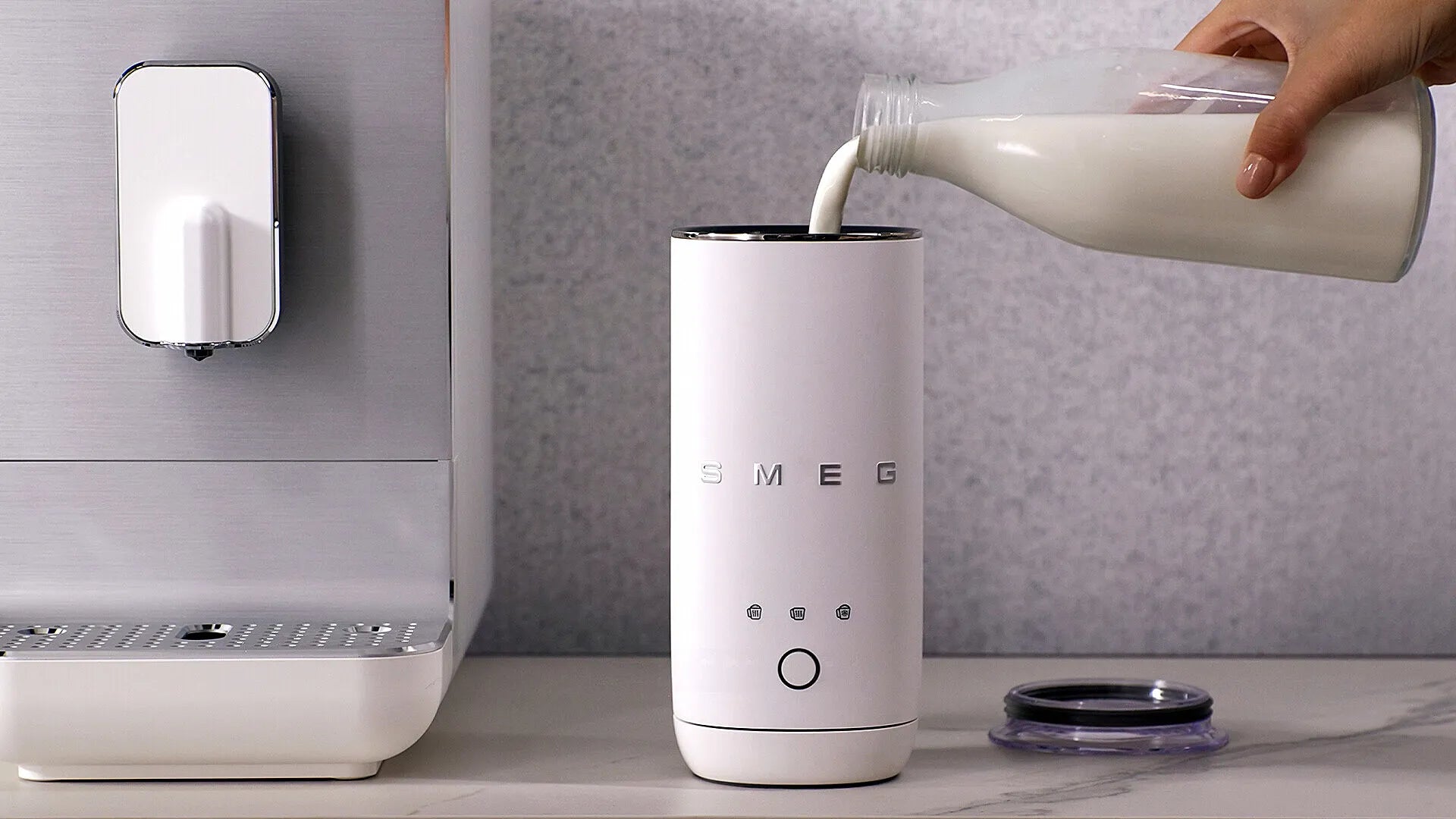 A white SMEG electric milk frother is shown on a countertop with milk being poured into its cup. A hand holds the milk bottle on the right, and a coffee machine is partially visible on the left against a gray textured wall.