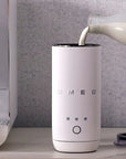 A white SMEG electric milk frother is shown on a countertop with milk being poured into its cup. A hand holds the milk bottle on the right, and a coffee machine is partially visible on the left against a gray textured wall.