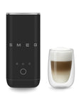 A black SMEG milk frother shown next to a double-walled glass cup filled with layered latte art, on a clean white background.