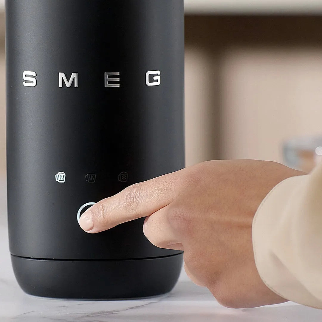 A close-up of a black SMEG milk frother with a hand pressing the central button, set against a softly blurred indoor background.