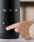 A close-up of a black SMEG milk frother with a hand pressing the central button, set against a softly blurred indoor background.