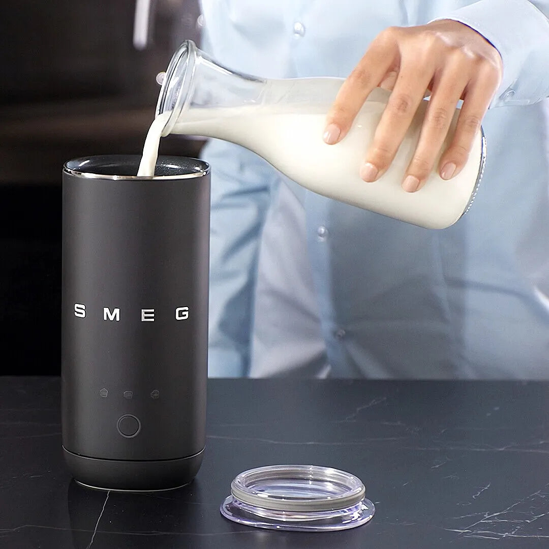 A person in a light blue shirt pours milk from a glass bottle into a black SMEG milk frother, with the clear lid placed beside it on a dark countertop.