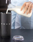 A person in a light blue shirt pours milk from a glass bottle into a black SMEG milk frother, with the clear lid placed beside it on a dark countertop.