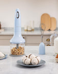 Handheld food chopper on a kitchen counter with various ingredients and kitchen tools.