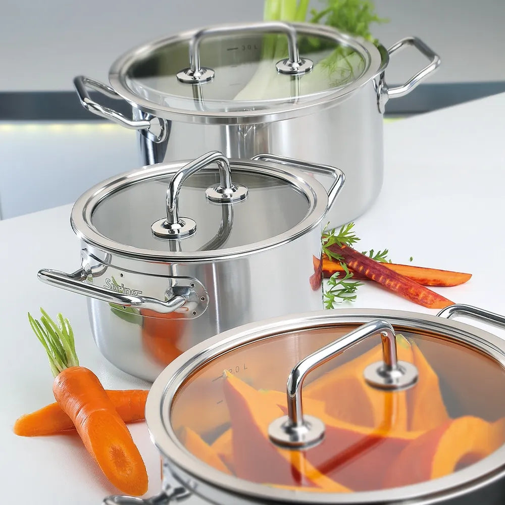 Three stainless steel stock pots with glass lids are arranged on a white countertop with fresh carrots and herbs as props.