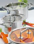 Three stainless steel stock pots with glass lids are arranged on a white countertop with fresh carrots and herbs as props.