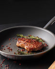 Pan with seared meat and herbs on a dark surface with vegetables and spices.