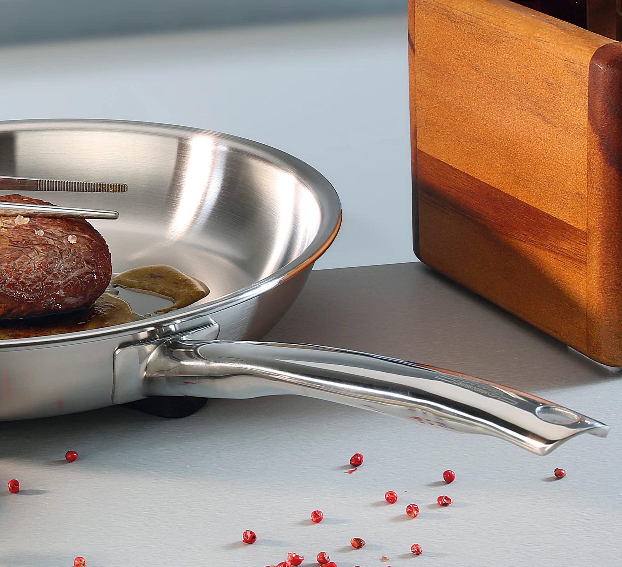 Stainless steel frying pan with a piece of meat on a light gray surface with red peppercorns.