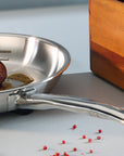 Stainless steel frying pan with a piece of meat on a light gray surface with red peppercorns.