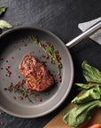Frying pan with meat and herbs on a dark surface with vegetables around