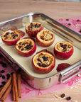 Stuffed apples in a baking dish on a pink floral tablecloth with cinnamon sticks and raisins.