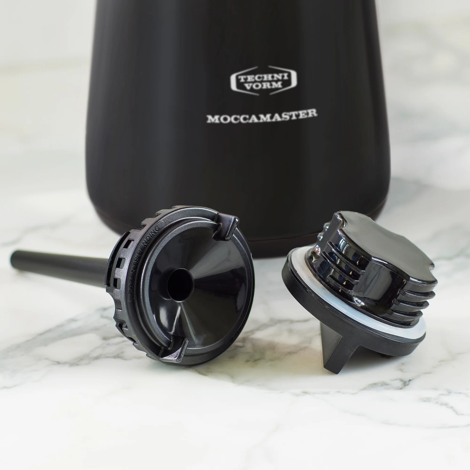 Two black coffee filters in front of a Moccamaster coffee pot on a marble surface.