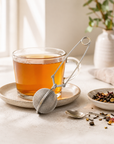 Little dipper tea infuser leaning against a clear, glass mug of tea next to tea leaves on a a granite table in a living room