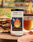 S'mores tea packaging with s'mores and a cup of tea on a wooden surface with a blurred campfire in the background.