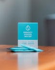 Box of Perfect Coffee Water with 4 pouches on a wooden surface and blurred light gray background