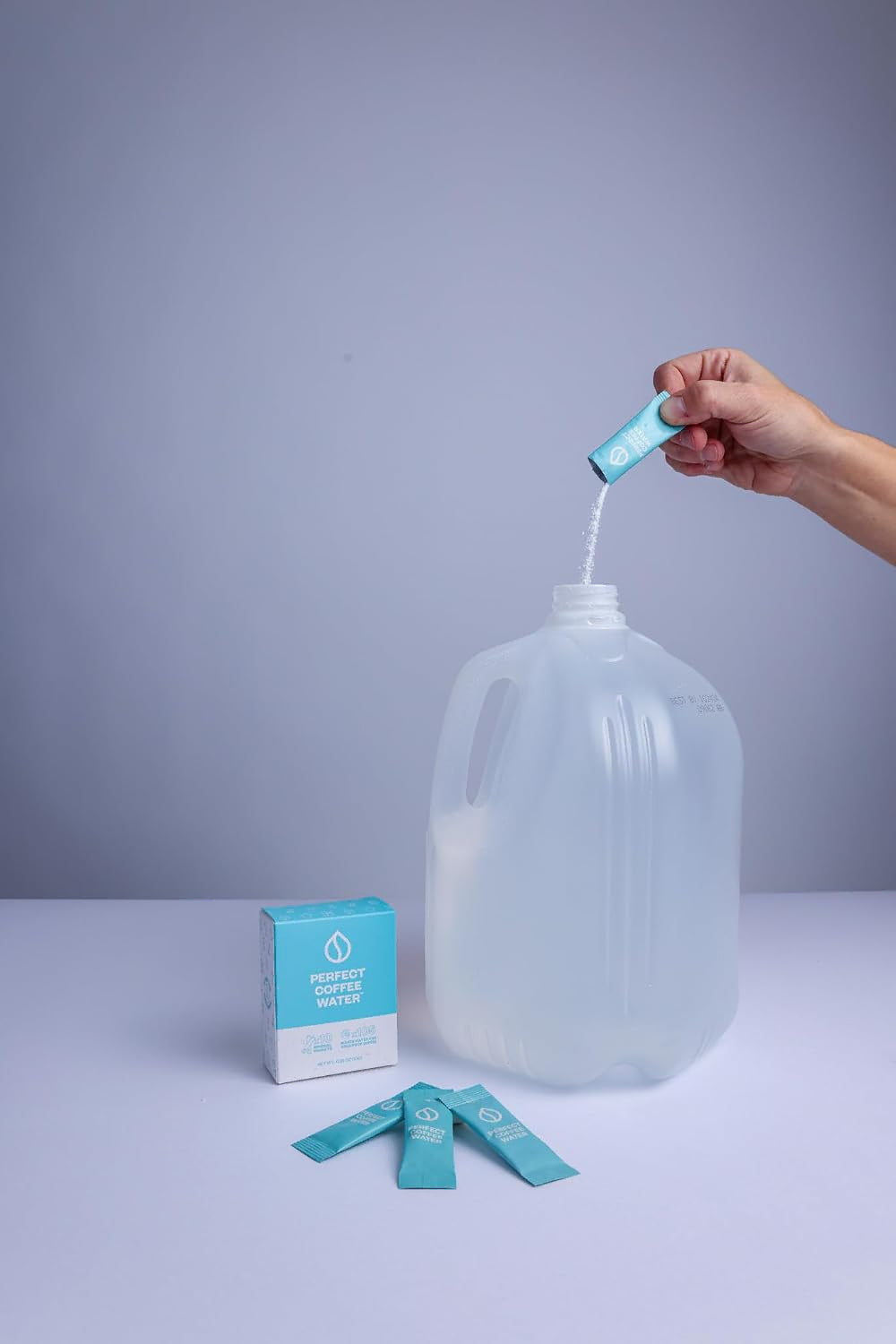 Someone pouring powder from pouch into a 1 gallon water jug next to 3 pouches and a packing displayed on a white surface and light gray background