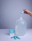 Someone pouring powder from pouch into a 1 gallon water jug next to 3 pouches and a packing displayed on a white surface and light gray background