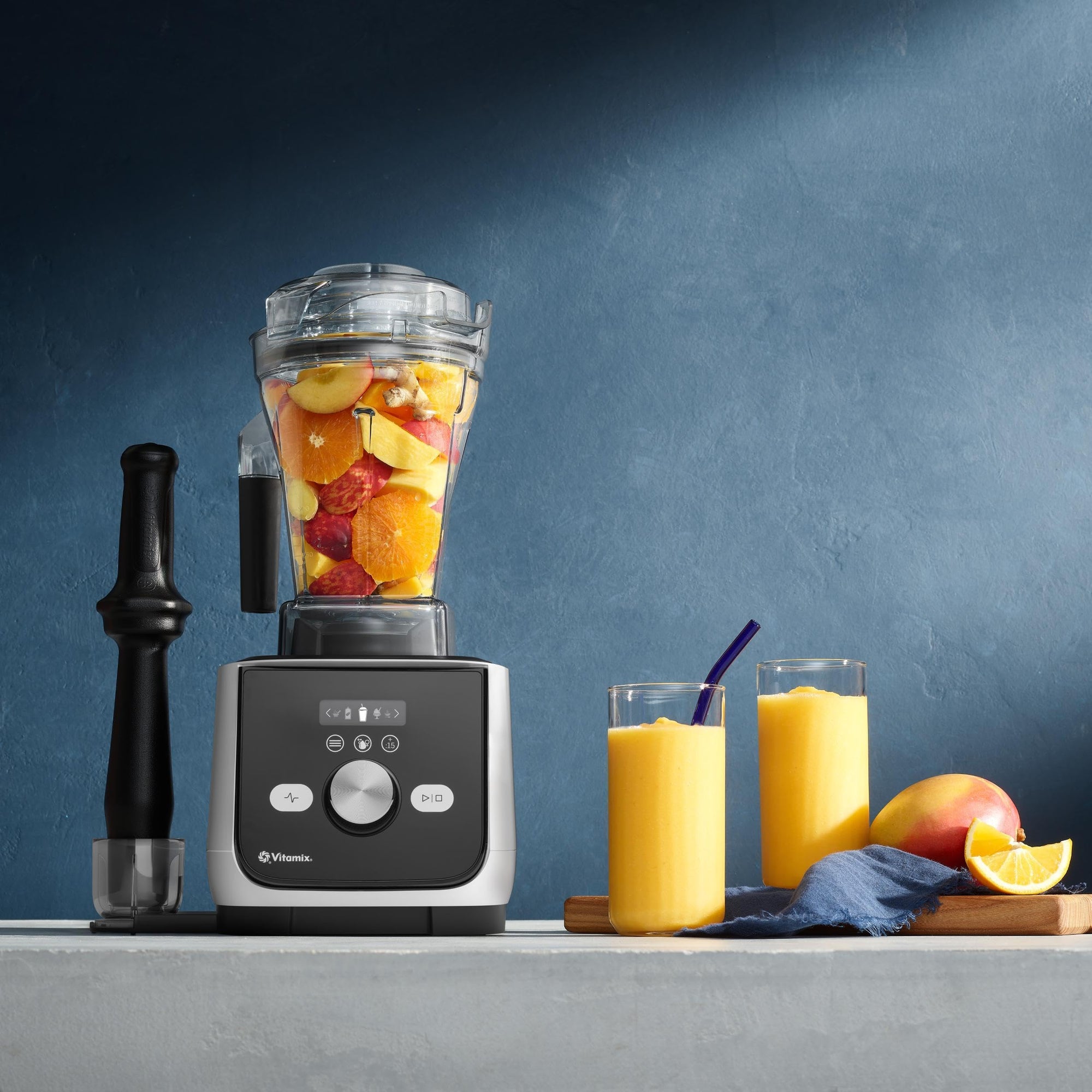 Blender with fruits, two glasses of juice, and a cutting board with a knife on a blue background