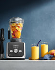 Blender with fruits, two glasses of juice, and a cutting board with a knife on a blue background